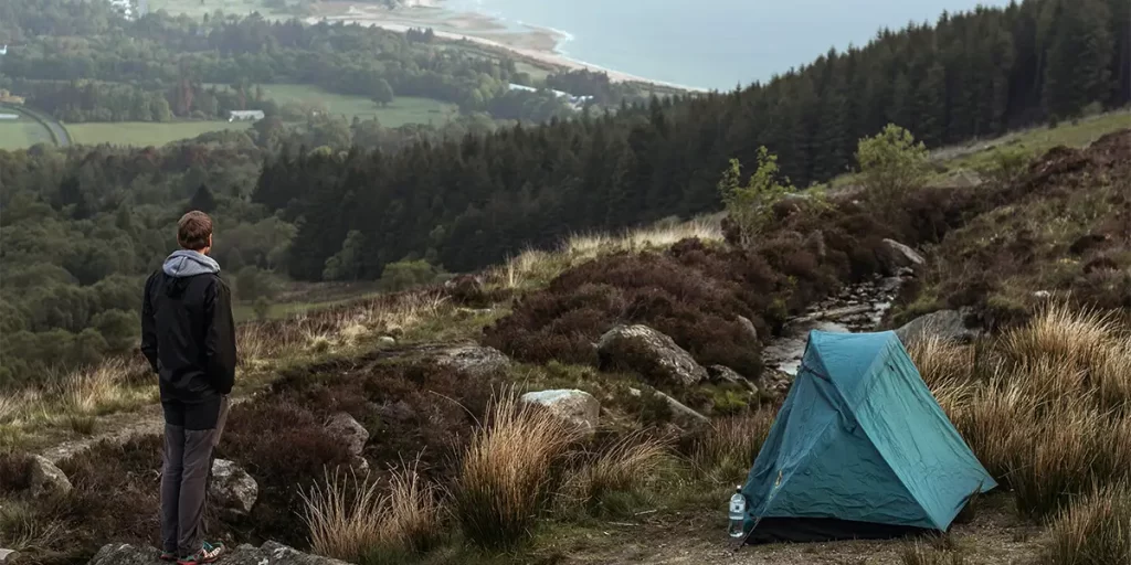 Campsite Selection: A hiker beside his ultralight backpacking tent, strategically pitched among the protective cover of bushes on the only relatively flat ground available