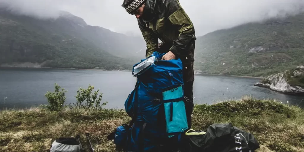Frameless vs. Framed Backpacks: Hiker sorting through his heavy framed backpack next to a lake between mountains