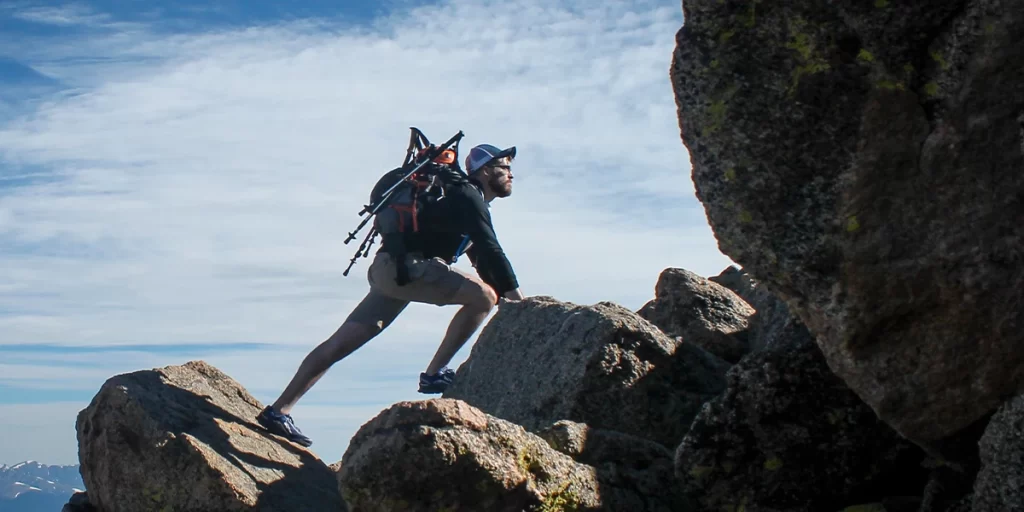 Attaching Hiking Poles: Hiker with hiking poles attached to his backpack climbing a steep rocky hill