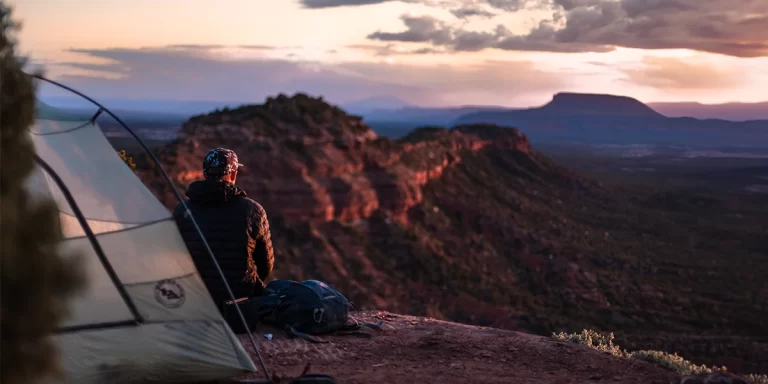 1-Person vs. 2-Person Tent: Hiker seated next to a 2-person tent at sunset in a picturesque mountain range