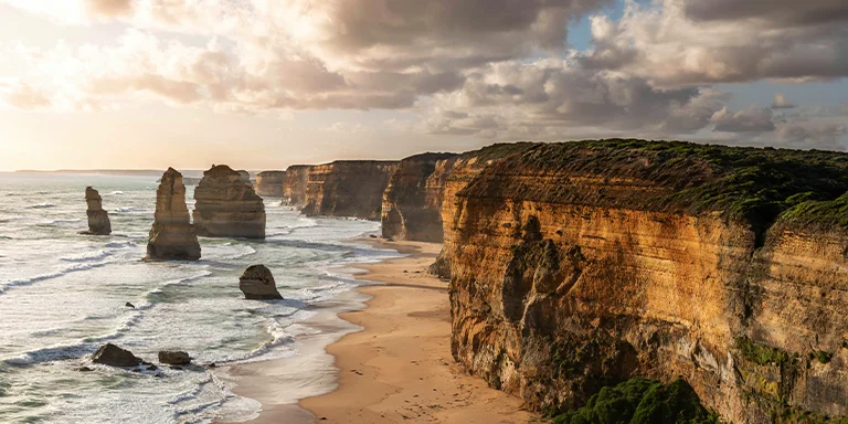 A sweeping beach in the foreground leads to towering sea cliffs bathed in the glow of sunset at the Twelve Apostles along Australia's Great Ocean Road