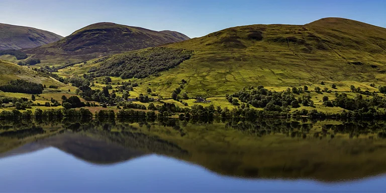 A serene green field stretches out to meet the calm, mirror-like waters of a scenic Scottish loch, surrounded by verdant hills