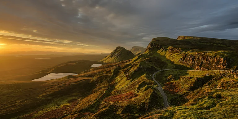 An aerial view from above shows a winding road traversing through the rugged, mountainous landscape of Scotland's Isle of Skye at sunset
