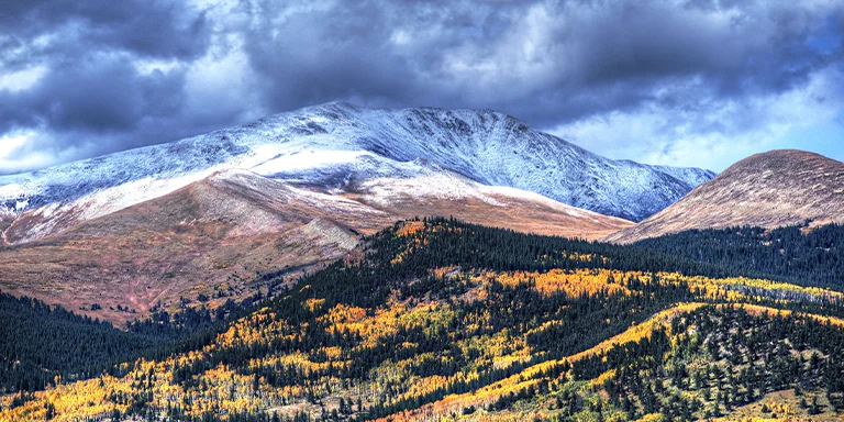 Dark storm clouds loom over a rugged mountain peak while bright fall foliage lines the highway, creating a moody landscape in this autumn image overlooking Kenosha Pass in Colorado