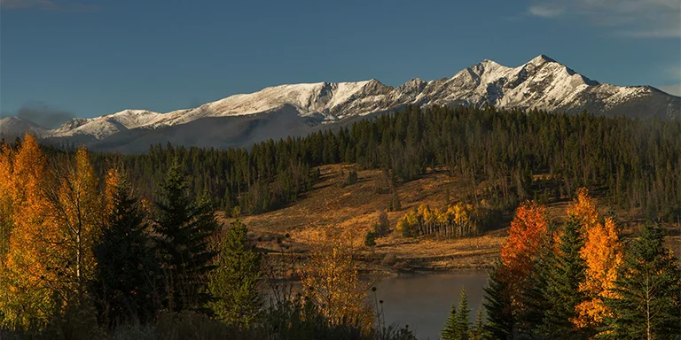 The majestic snow-capped peaks of the Rocky Mountains tower behind a foreground of vibrant green pine trees and golden aspens in this beautiful daytime image of Breckenridge, Colorado