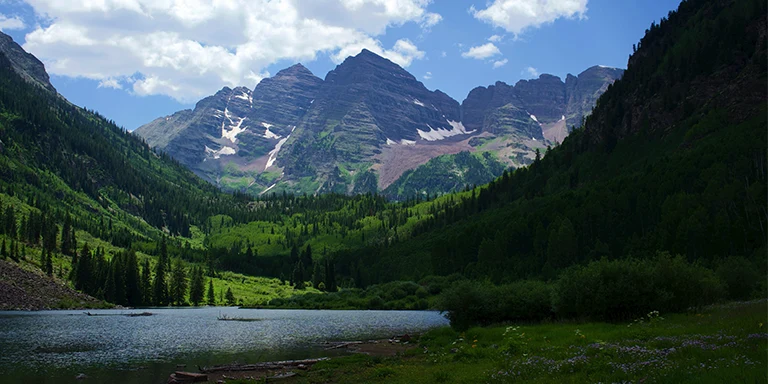 This scenic photo captures the breathtaking natural splendor of Colorado's Maroon Bells, with verdant evergreen trees framing the symmetrical snow-dusted peaks reflected in the calm waters of a mountain lake