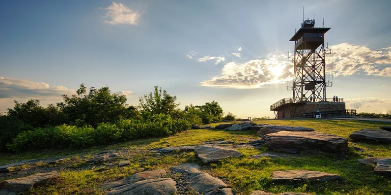 The worn observation tower stands tall on the bare rocky summit of Mount Wachusett against a clear blue sky, with green forests