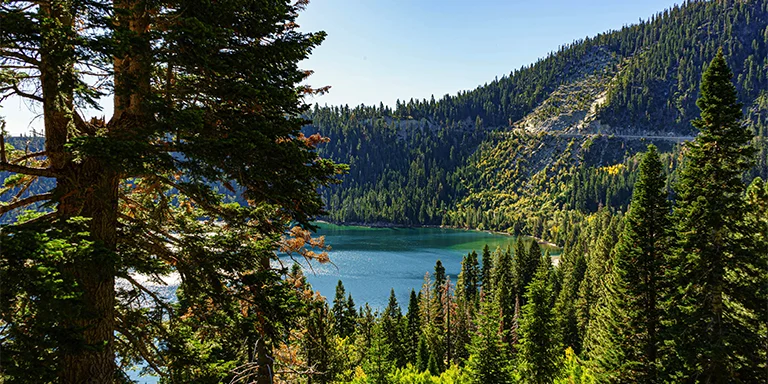 The photo shows a serene mountain lake surrounded by evergreen trees with snow-capped peaks in the background