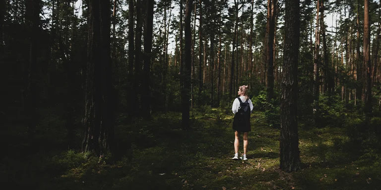 A woman traveler wearing a white long sleeve shirt and black skirt stands surrounded by tall trees and lush green foliage while exploring Kampinos Forest in Poland on a sunny day