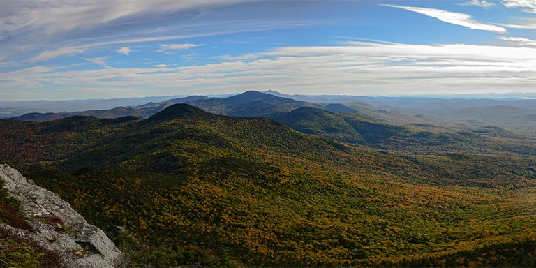 A panoramic summit view of the green mountains in Vermont from Mount Mansfield, with dense coniferous forests blanketing the rolling peaks and valleys below a bright blue sky