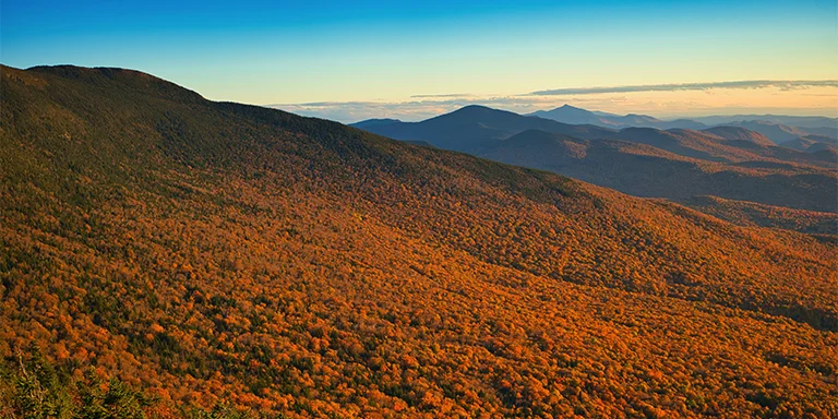 A landscape image of Mount Mansfield's brilliant fall foliage in Stowe, Vermont, with orange, red, and yellow trees covering rolling green hills under a bright blue sky