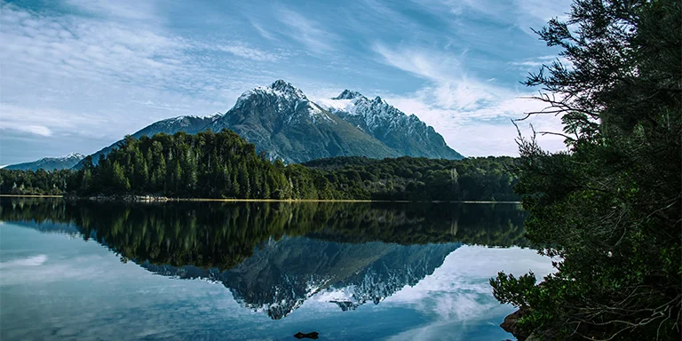 Misty blue waters and dark evergreens encircle the shore beneath towering peaks in this tranquil woodland scene by an alpine lake in Bariloche