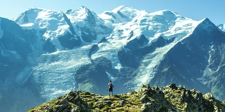 A lone hiker gazes out at the dramatic panorama of snow-capped mountain peaks stretching across the horizon under a crisp blue sky, taken atop a grassy hill