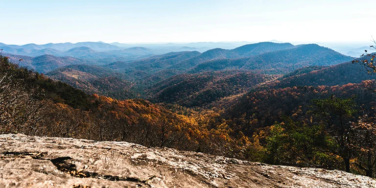 A sweeping vista overlooks forested mountains blanketed in fall foliage below a bright blue sky