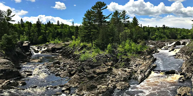 This serene scene from Jay Cooke State Park in Carlton, Minnesota captures the lush green foliage lining the banks of a peaceful river under a brilliant blue sky