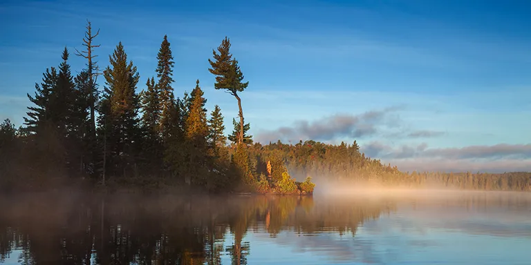 Shrouded in ethereal morning fog, this tranquil sunrise view showcases the glassy blue waters of a northern Minnesota lake reflecting the changing colors of an island's trees in early September
