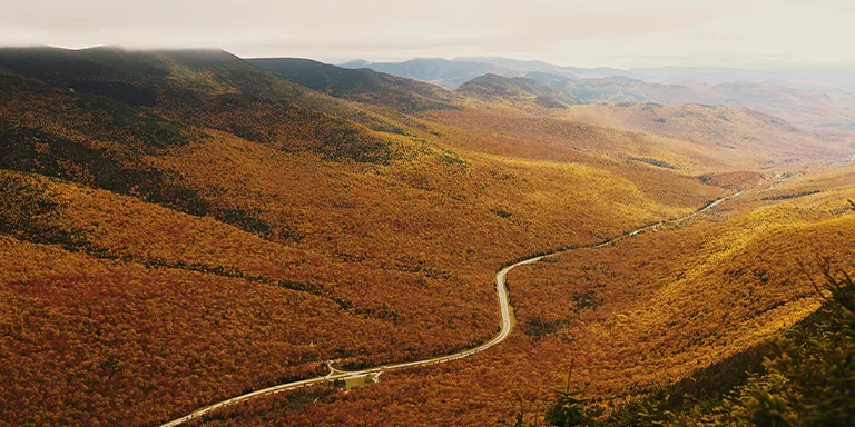 This aerial view captures the rugged, tree-covered peaks and ridges of the White Mountain National Forest near Livermore, New Hampshire under a bright daytime sky