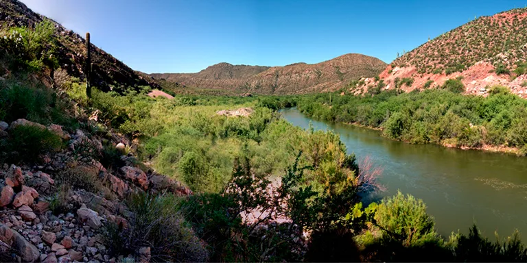 The Gila River flows swiftly over rocky terrain just downstream from the towering Coolidge Dam, its waters sparkling in the sunlight