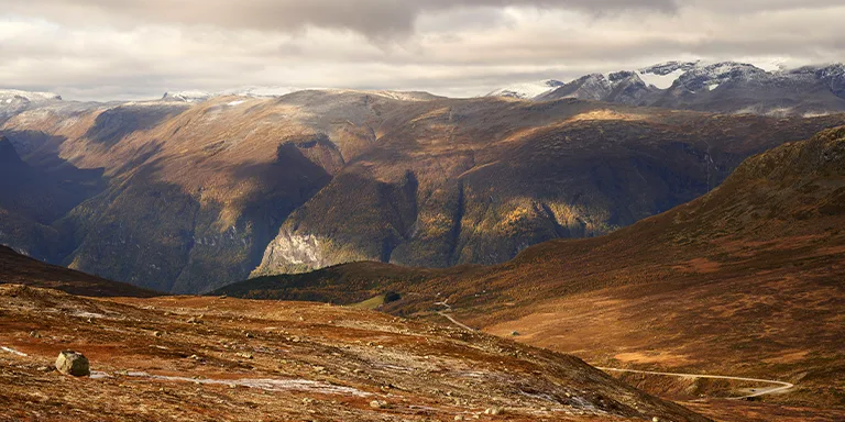 A towering mountain range extends across a fjord landscape, with snowcapped peaks in the distant backdrop framed by changing autumn foliage in the foreground