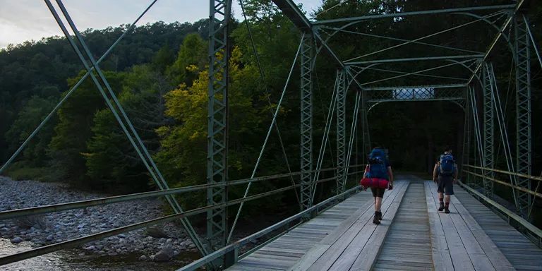 Two backpackers cross a rustic bridge spanning a river, immersed in the tranquil beauty of Pennsylvania's forests during their outdoor adventure