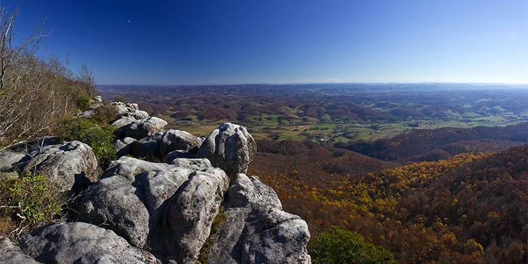 A vista showing a sunny valley blanketed with trees and foliage below towering rock formations along the trail at White Rocks in Cumberland Gap National Historic Park, United States