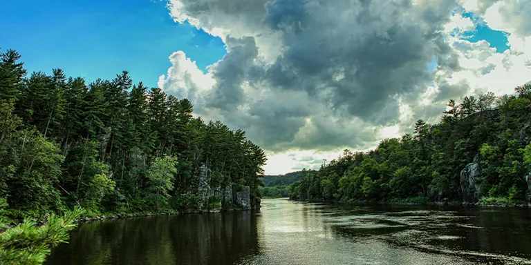 A scenic view of lush green trees lining a river under a clear blue sky with white clouds at Wisconsin Interstate State Park

