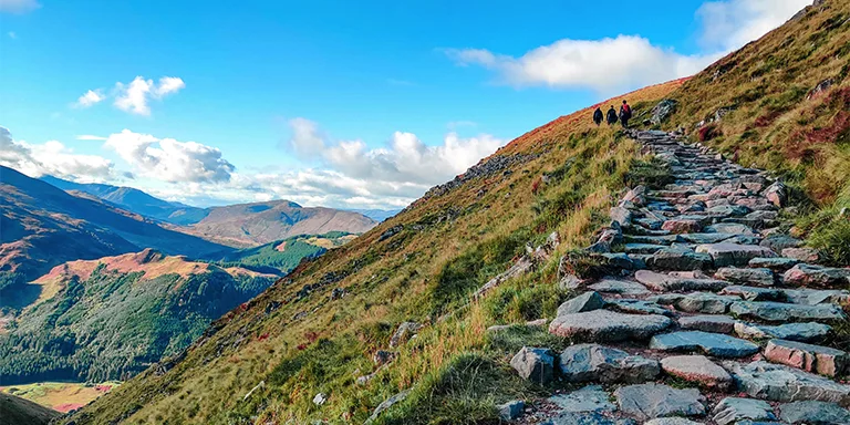 A winding gray rocky path leads up a grassy slope towards the majestic snow-capped peak of Ben Nevis mountain against a brilliant blue sky