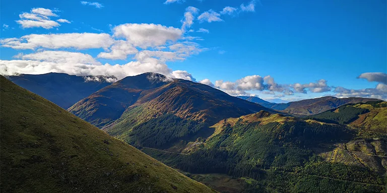 A rugged green and brown mountain peak rises towards a brilliant blue sky, capturing the scenic beauty of a hiking trail on Ben Nevis in Scotland