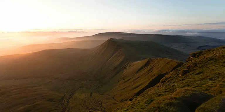 A breathtaking aerial view of a mountainous range bathed in the warm golden light of the sunrise or sunset, captured from the summit of Pen y Fan in the Brecon Beacons, United Kingdom
